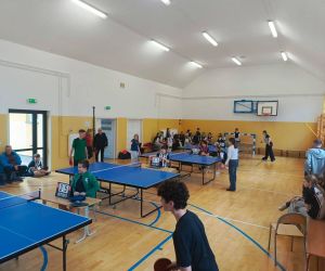 Zawodnicy rozgrywający mecze tenisa stołowego w sali gimnastycznej, widoczni sędziowie i kibice.