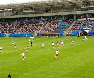 Fragment meczu piłkarskiego widziany z trybun, zawodnicy w trakcie gry na boisku.