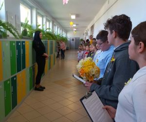 Uczniowie ustawieni na szkolnym korytarzu podczas apelu, część z nich trzyma żonkile i kartki z tekstem.