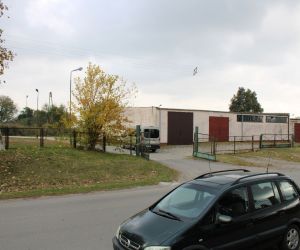 Wjazd na plac od drogi powiatowej z widoczną częścią budynku magazynu.