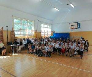 Uczniowie siedzą na ławkach, a w tle nauczyciele
