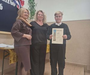 Uczeń prezentuje dyplom zdobyty w konkursie, towarzyszą mu nauczycielki podczas uroczystego wręczenia nagród.