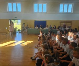 Pani Dyrektor przemawia podczas uroczystego rozpoczęcia zakończenia roku szkolnego. Na sali gimnastycznej zgromadzeni są uczniowie, nauczyciele oraz rodzice.