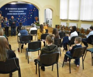 Grupa ludzi wewnątrz budynku siedzi na krzesłach, przy scenie znajdują się dwie osoby.