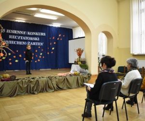 Grupa ludzi wewnątrz budynku siedzi na krzesłach, dziecko występuje na scenie