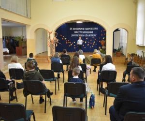 Grupa ludzi wewnątrz budynku siedzi na krzesłach, dziecko wystepuje na scenie.