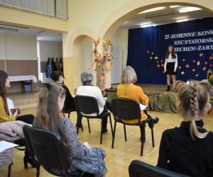 Grupa ludzi wewnątrz budynku siedzi na krzesłach, dziecko występuje na scenie.