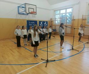 Uczniowie prezentują program artystyczny przed zgromadzoną publicznością. Kilkoro z nich stoi przy mikrofonach, a pozostali tworzą grupę występujących.