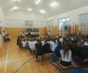 Widok na salę gimnastyczną podczas obchodów Dnia Edukacji Narodowej. Na pierwszym planie uczniowie prezentują część artystyczną, a na ławkach siedzą licznie zgromadzeni uczniowie, nauczyciele i pozostali uczestnicy uroczystości.