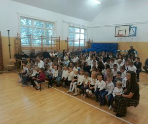 Najmłodsze dzieci, starsi uczniowie oraz nauczyciele zgromadzeni na sali gimnastycznej podczas szkolnych obchodów Dnia Edukacji Narodowej. Zdjęcie przedstawia publiczność uczestniczącą we wspólnym świętowaniu.