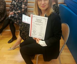 Nauczycielka siedząca na krześle z uśmiechem prezentuje pamiątkowe „Świadectwo Najlepszej Wychowawczyni”.