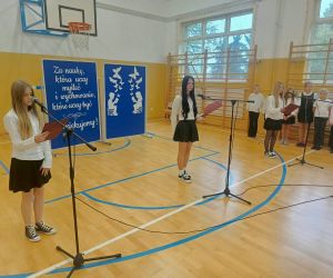 Uczennice prowadzą uroczystą akademię z okazji Dnia Edukacji Narodowej. Przy mikrofonach odczytują przygotowane teksty z czerwonych teczek. W tle pozostali uczniowie oczekują na swoją część występu.