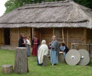 Kobiety w antycznych strojach na tle drewnianej chaty, pokrytej strzechą.
