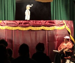 Kukiełki w przedstawieniu teatrzyku kukiełkowym. Narrator siedzi przed parawanem.