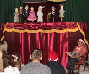 Kukiełki w przedstawieniu teatrzyku kukiełkowym. Narrator siedzi przed parawanem. Widać publiczność siedzącą tyłem.