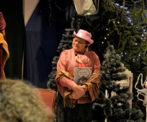 Narrator przedstawienia teatralnego stoi przed parawanem, wewnątrz budynku..