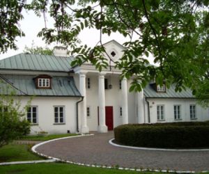 Biały dwukondygnacyjny budynek z zielonym dachem, Przed wejściem stoją cztery kolumny sięgające wyższej kondygnacji. Na dziedzińcu przed obiektem żywopłot a dookoła droga/chodnik