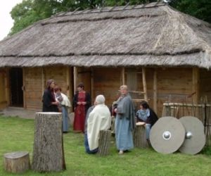 Ludzie w kolorowych szatach  stoją przed drewnianą chatą pokrytą strzechą.