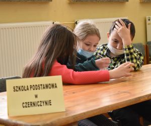 Dzieci siedzą wewnątrz budynku i rozwiązują zadania konkursowe.