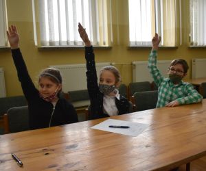 Dzieci siedzą wewnątrz budynku i trzymają w górze ręce.