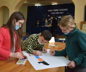 Dzieci siedzą wewnątrz budynku i rozwiązują zadania konkursowe.