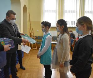 Dzieci i osoby dorosłe wewnątrz budynku. Mężczyzna wręcza nagrody dyplom.