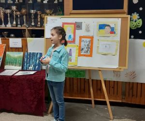 Dziewczynka ubrana w niebieską kurtkę układa kostkę rubika, w tle stoi tablica z pracami plastycznymi innych uczestników.