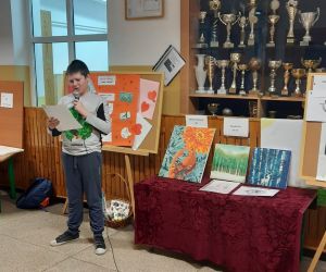 Chłopiec w szarej bluzie prezentuje nowinki motoryzacyjne, omawia min. budowę i zasadę działania silnika w modelach marki Bmw. Obok i za nim znajdują się prace plastyczne innych uczniów.