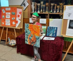 Dziewczynka w zielonej czapeczce trzyma przed sobą obraz wykonany metodą malowania po numerach. W tle , na stoliku znajdują się prace innych uczestników.