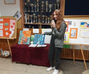 Dziewczynka ze starszej klasy prezentuje swój dorobek plastyczny. Wśród prac są portrety i pejzaże wykonane ołówkiem i farbami. 