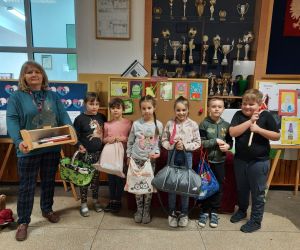 Nauczycielka i uczniowie stoją na korytarzu, kobieta trzyma w ręku drewniany chlebak, uczniowie zamiast plecaków mają worek na kiju, torbę sportową, reklamówki, koszyk zakupowy.