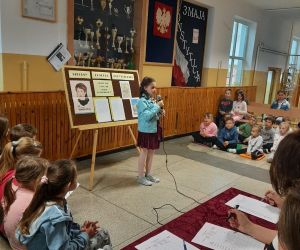 Dziewczynka w niebieskiej kurtce recytuje wiersz. Uczniowie siedzą i słuchają recytacji a jury zastanawia się nad punktacją.