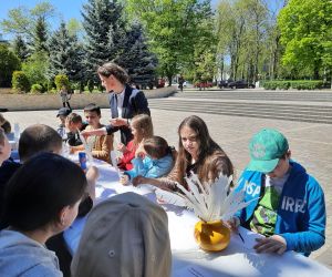 Pani bibliotekarka rozdaje uczniom gęsie pióra oraz podaje tusz i kartki. Uczniowie siedzą przy stoliku i przepisują tekst gęsim piórem.