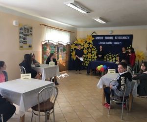 Trzyosobowe drużyny uczniów przy stolikach. W głębi trzy kobiety prowadzące turniej, na tle tablicy z nazwą turnieju. Tablica udekorowana żółtymi kwiatami.