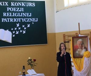 Uczennica w granatowej sukience recytuje. W tle portret Papieża Polaka.