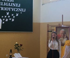 Uczennica w biało-czarnym stroju recytuje. W tle portret Papieża Polaka.