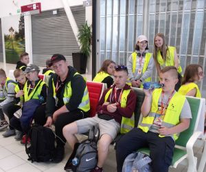 Uczniowie siedzący i stojący w  poczekalni do odlotów statkami powietrznymi