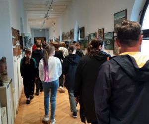 Uczniowie i opiekunowie przemieszczają się korytarzem w muzeum, na ścianach wiszą obrazy a po bokach stoją rzeźby