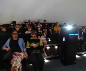 Uczniowie z opiekunami w planetarium w Ogrodzie Doświadczeń w Krakowie. Siedzą w fotelach w oczekiwaniu na projekcje filmu 3D.