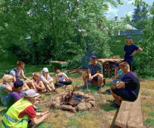 Grupa osób piecze kiełbaski przy ognisku.