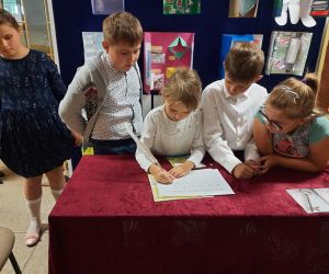 Uczniowie stoją przy stole, jedna z uczennic przepisuje tekst z bajki