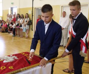 Uczeń klasy ósmej wbija pamiątkowy gwóźdź w drzewce sztandaru, a drugi uczeń przytrzymuje sztandar