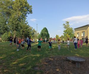 Na zdjęciu widoczna jest grupa dzieci trzymających się za ręce w trakcie zabawy na świeżym powietrzu.