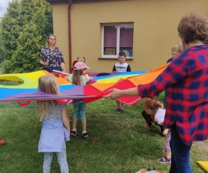 Zdjęcie zrobione na zewnątrz przed budynkiem, znajdują się na nim dzieci i panie opiekunki w trakcie zabawy chusta animacyjną.