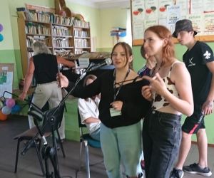 Na zdjęciu znajduje się pięć osób, dwie z nich stoją przy mikrofonie.