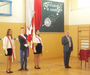 Pan dyrektor wygłasza przemowę. W tle poczet sztandarowy – chłopiec trzyma sztandar obok dwie dziewczynki.