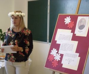 Nauczycielka ubrana w czarną kwiecistą bluzkę i wianek z kwiatów na głowie przy tablicy poświęconej Adamowi Mickiewiczowi