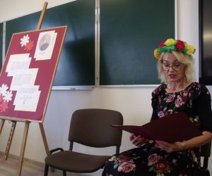 Nauczycielka w czarnej kwiecistej sukience, z wiankiem z kolorowych kwiatów na głowie, czyta tekst ballady