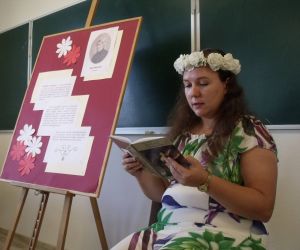 Nauczycielka w białej, kwiecistej sukience i w wianku z kwiatów na głowie, czytająca tekst ballady
