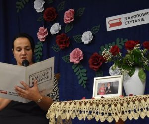 Zdjęcie przedstawia kobietę siedzącą przy stoliku, która czyta z kartki.